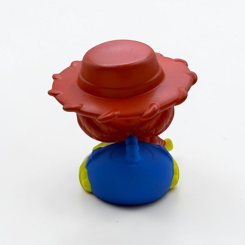 K&ouml;nnte beinhalten: Eine kleine Figur mit einem roten Cowboyhut mit breiter Krempe, der &uuml;ber einem Kopf mit rotem, geflochtenem Haar sitzt. Die Figur tr&auml;gt ein leuchtend blaues Hemd mit gelben Akzenten an den Seiten, betrachtet von hinten vor wei&szlig;em Hintergrund.