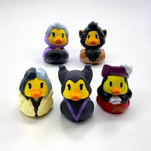 Puede incluir: Colección de seis patitos de goma, cada uno diseñado para parecerse a un villano de Disney diferente. Los patos presentan colores vibrantes y disfraces detallados, incluyendo personajes como Cruella de Vil y Maléfica. Los patos son amarillos con picos naranjas.
