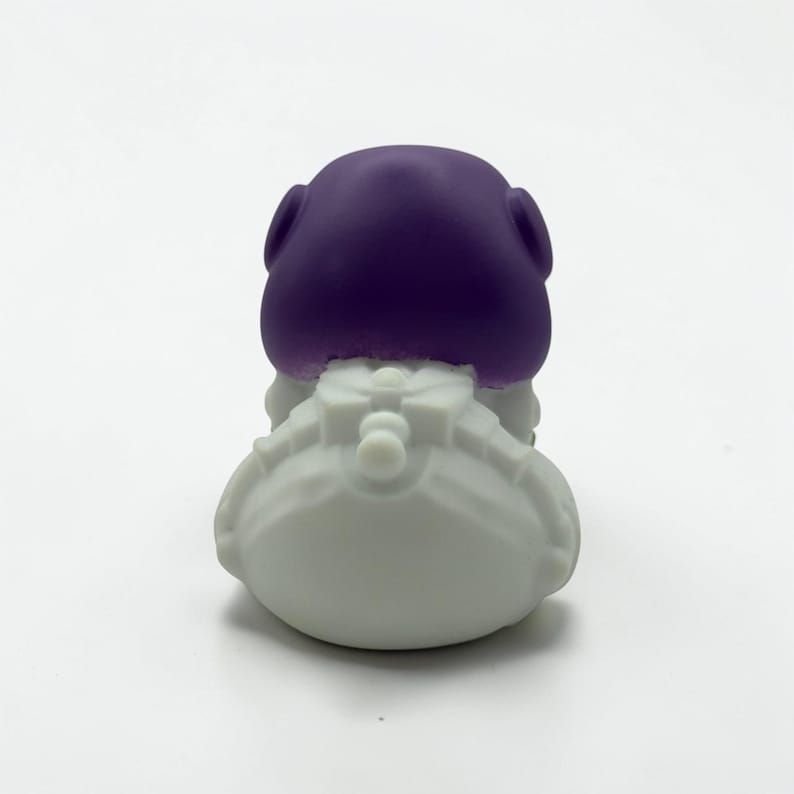 K&ouml;nnte beinhalten: Eine kleine Figur mit einem glatten, abgerundeten violetten Kopf auf einem wei&szlig;en, strukturierten K&ouml;rper. Der wei&szlig;e Bereich zeigt geformte Details, die an einen Rucksack oder Ausr&uuml;stung erinnern, mit einem zentralen runden Element und Segmenten.
