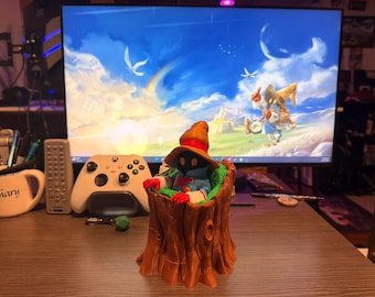 May include: A Final Fantasy IX Vivi Ornitier figurine in a tree stump. The figure is wearing a black robe, red gloves, and a yellow hat. The background includes a computer monitor with a Final Fantasy IX wallpaper, a white Xbox controller, and a mug.