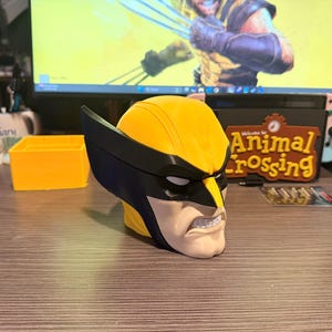 May include: A 3D printed bust of the character Wolverine featuring a bright yellow mask with black accents, white eye slits, and a tan lower face showing a gritted teeth expression. The head sculpture displays sharp black ear fins and a smooth finish.