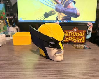 May include: A 3D printed bust of the character Wolverine featuring a bright yellow mask with black accents, white eye slits, and a tan lower face showing a gritted teeth expression. The head sculpture displays sharp black ear fins and a smooth finish.
