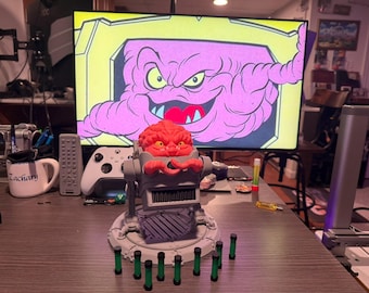 May include: A detailed action figure of Krang from Teenage Mutant Ninja Turtles, featuring a red brain-like head and a robotic body. The figure is gray and red, with green cylindrical accessories. A large screen displays a cartoon image of Krang.