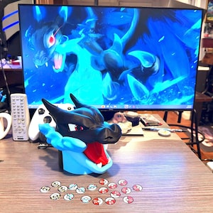 May include: A black, red, and light blue Charizard-themed figurine with an open mouth, and a set of small, round, colourful game tokens. A computer monitor displays a blue dragon graphic.