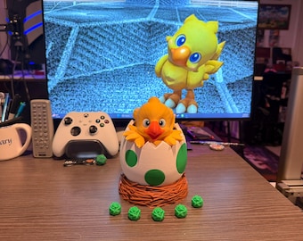 May include: A yellow and orange Chocobo figurine emerging from a white egg with green spots, set in a brown nest. A larger Chocobo figure stands behind it. Several small green rose-shaped decorations are in front of the egg. A white Xbox controller is visible.