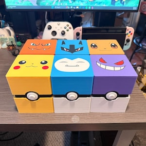 Könnte beinhalten: Sechs farbenfrohe, würfelförmige Aufbewahrungsboxen mit Gesichtern von Pokémon-Charakteren auf den Deckeln. Die Boxen sind gelb, blau, orange und lila, mit weißen und schwarzen Akzenten. Jede Box hat ein weißes und schwarzes kreisförmiges Design auf der Vorderseite, das einem Pokéball ähnelt.