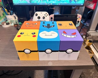 May include: Six colorful, cube-shaped storage boxes with Pokémon character faces on the lids. The boxes are yellow, blue, orange, and purple, with white and black accents. Each box has a white and black circular design on the front, resembling a Poké Ball.