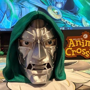 Op de afbeelding: Een 3D-geprint Doctor Doom-masker met een metallic zilveren afwerking, zichtbare klinknageldetails en een textuurrijke groene kap. Het masker toont een strenge gezichtsuitdrukking met zichtbare ogen en een gestructureerde kaaklijn.