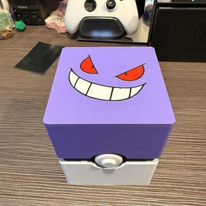May include: A purple and white box with a Gengar face design on top. The box is a cube shape with a white base and a purple top. The Gengar face has red eyes and a wide grin. The box is designed to look like a Pokeball.
