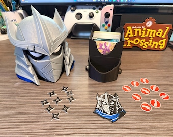 May include: A gray and blue Shredder helmet-shaped card holder, a black card holder, ninja stars, and game tokens. The card holder has a slot for cards and a small compartment. The game tokens are red and white.