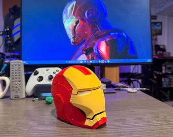 May include: A red and yellow Iron Man helmet replica, with a yellow faceplate and red accents. The helmet is sitting on a wooden surface, with a computer monitor displaying an Iron Man image in the background. A white game controller and remote are also visible.