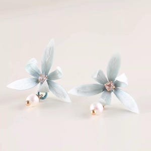 May include: A pair of pale blue flower earrings with a pearl drop. Each earring has a five-petal flower design with a small crystal in the centre. The earrings are set against a soft, neutral background.