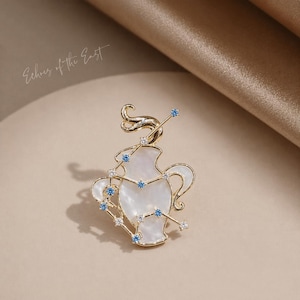 May include: Gold-toned brooch in the shape of a vase, adorned with a white mother-of-pearl inlay and blue and clear crystal accents. The brooch features a constellation design and the text "Echoes of the East".
