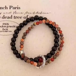 May include: Two beaded bracelets. One bracelet features black beads, the other has a mix of red, brown, and orange beads. The bracelets are connected by a silver-coloured clasp. The background has text.