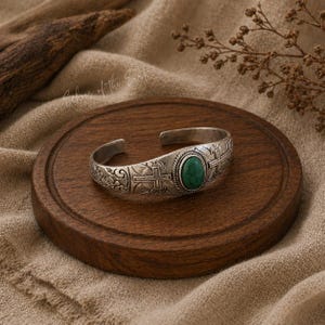 May include: Silver cuff bracelet with an oval, green gemstone centre. The bracelet features a cross design and floral patterns. The jewellery is displayed on a round, wooden tray.