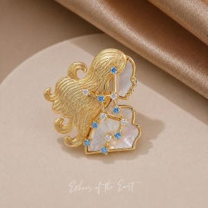 May include: Gold-toned brooch featuring a woman's profile and flowing hair, adorned with blue and clear gemstones. The design incorporates a constellation pattern on a mother-of-pearl background. The text "Echoes of the East" is at the bottom.