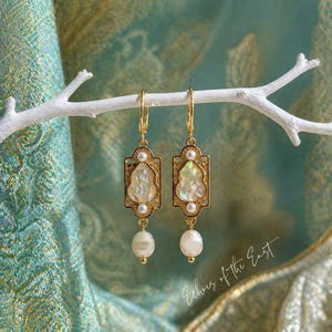 Può includere: Un paio di orecchini color oro con un design rettangolare, con centri iridescenti, piccole perle e perle pendenti più grandi. Gli orecchini sono sospesi a un ramo bianco su uno sfondo turchese e oro. Il testo "Echoes of the East" è visibile.