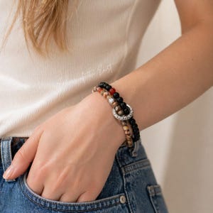 May include: Two beaded bracelets with a mix of earth-toned and black beads. One bracelet features a silver-colored clasp. The bracelets are worn on a wrist, with the hand in a denim pocket. The bracelets are a stylish accessory.