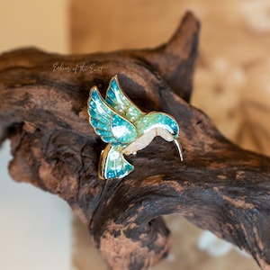 May include: A gold-coloured hummingbird brooch with turquoise and green enamel wings and body. The bird is perched on a piece of dark, textured wood. The brooch has a detailed design and a long, slender beak.