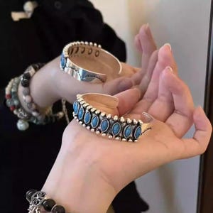 Könnte beinhalten: Zwei silberfarbene Armreifen mit ovalen, türkisfarbenen Steinen. Die Armreifen haben ein detailliertes Design mit kleinen silbernen Perlen. Die offenen Armreifen werden auf den Händen einer Person präsentiert.