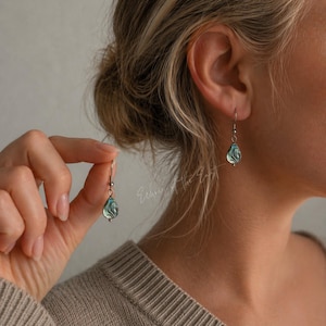 Könnte beinhalten: Silberne Hängeohrringe mit tropfenförmigen, irisierenden blau-grünen Anhängern. Die Ohrringe werden am Ohr präsentiert, um das Design und die Farbe hervorzuheben. Der Schmuck hat ein schlichtes, elegantes Design.