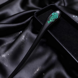 May include: A black wooden hair stick with a bright green Celtic knot design at the top. The hair accessory is presented in a black box on a black satin surface.