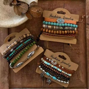 Könnte beinhalten: Eine Auswahl an gestapelten Armbändern auf braunen Pappkarten. Die Armbänder bestehen aus verschiedenen Materialien, darunter Perlen in Türkis, Braun und Grün, Leder und Metallakzenten. Auf den Karten steht "Fashion Jewelry".