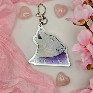 Könnte beinhalten: Ein silberfarbener Schlüsselanhänger mit einem heulenden Wolfsdesign. Der Wolf ist mit einem lila Umhang dargestellt, der mit Monden und Sternen verziert ist. Der Schlüsselanhänger ist an einem silbernen Verschluss befestigt. Rosa herzförmige Steine und rosa Blumen umgeben den Schlüsselanhänger.