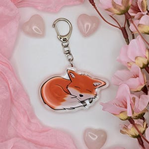 Könnte beinhalten: Ein klarer Acryl-Schlüsselanhänger mit einer Illustration eines schlafenden Fuchses in Orange und Weiß. Er hat einen silberfarbenen Verschluss und eine Kette. Der Hintergrund umfasst rosa Stoff, herzförmige Steine und rosa Blumen.
