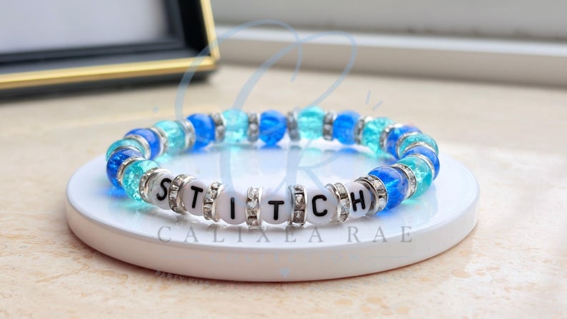 May include: A beaded bracelet with the word "STITCH" spelled out in white letter beads. The bracelet features a mix of blue and clear beads, with silver accents. The bracelet is displayed on a white circular stand.