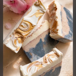 May include: Three rectangular bars of handcrafted soap featuring a layered design with a dark charcoal grey base, a light beige middle section, and a white top layer swirled with metallic gold accents, accompanied by light pink flower petals.