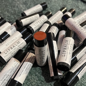 Peut inclure: Plusieurs tubes de baume à lèvres avec des bouchons noirs et des étiquettes blanches. Les étiquettes indiquent "LIPBALM", les ingrédients et les informations sur la marque. Un tube est ouvert, révélant un baume brun-rouge. Les produits sont sur une surface verte.