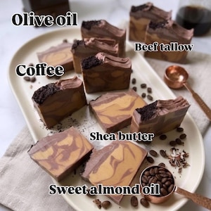 May include: Several rectangular bars of soap with a marbled brown and tan pattern, topped with dark coffee grounds. The text labels Cocoa, Olive oil, Beef tallow, Coffee, Shea butter, and Sweet almond oil are overlaid on the image.