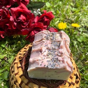 May include: A handmade soap bar, pale pink in colour, decorated with dried lavender. The soap sits on a small woven basket. Red roses and green grass form the background of the image.