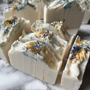 May include: Several rectangular bars of off-white soap arranged in a group. Each bar features a smooth, creamy texture with swirls of light blue colour and is topped with a scattering of dried yellow and blue flower petals on the top edge.
