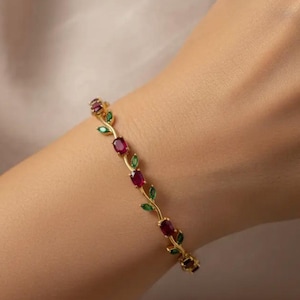 Peut inclure: Bracelet doré orné d'un motif de vigne avec des pierres rouges ovales et des feuilles vertes. Le bracelet est porté au poignet, mettant en valeur son esthétique délicate et élégante. Un bijou.