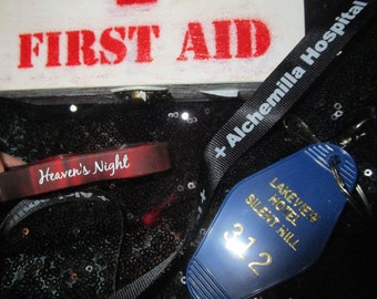 May include: A first aid kit with red text, a red wristband with "Heaven's Night" in white, a black lanyard with "+ Alchemilla Hospital +", and a blue hotel key tag with gold text that reads "Lakeview Hotel Silent Hill 312".