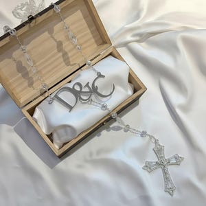 Puede incluir: Un rosario de tono plateado con cuentas transparentes facetadas, un colgante central de metal con las letras D&C en escritura estilizada y un crucifijo grande y ornamentado con cristales transparentes, presentado dentro de una caja de madera.