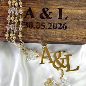 May include: A wooden box with the initials "A & L" and the date "30.05.2026" engraved on the front. A gold-colored rosary with clear crystal beads and a cross is draped over the box. The rosary also has the initials "A & L".