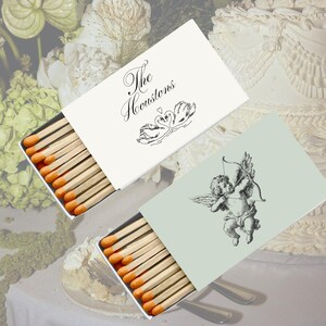 May include: Two rectangular matchboxes with orange-tipped wooden matches. One white box features the text The Houstons above a black line drawing of two swans. The other light green box features a black line drawing of a winged cherub with a bow.