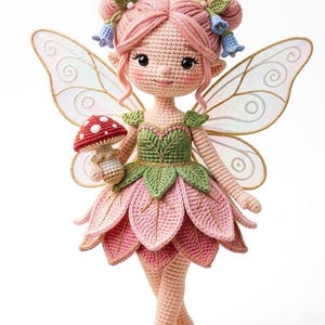 Op de afbeelding: Een handgemaakte gehaakte feeënpop met roze haar in knotjes, met een paddenstoel. De fee draagt een roze en groene jurk met bladvormige details en gouden accenten. Delicate, transparante vleugels en blauwe bloemaccenten maken het ontwerp compleet. De pop staat op een houten voet.