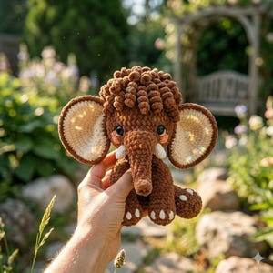 A person holds up a small brown crocheted mammoth toy outdoors with a garden backdrop.