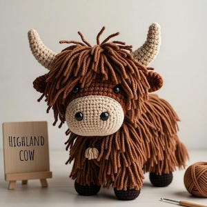 May include: A handmade crochet Highland cow with brown shaggy fur, beige face, and horns. The cow has black eyes and is standing on four black legs. A small wooden sign reads "Highland Cow".