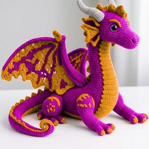 Puede incluir: Un dragón de ganchillo hecho a mano en morado y dorado. El dragón presenta alas, cuernos y una larga cola detalladas. Un artículo artesanal y caprichoso para los entusiastas de la fantasía.
