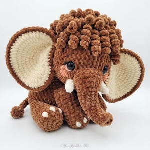 A crochet brown mammoth amigurumi toy sitting on a clean white surface with a soft, curly head mane.