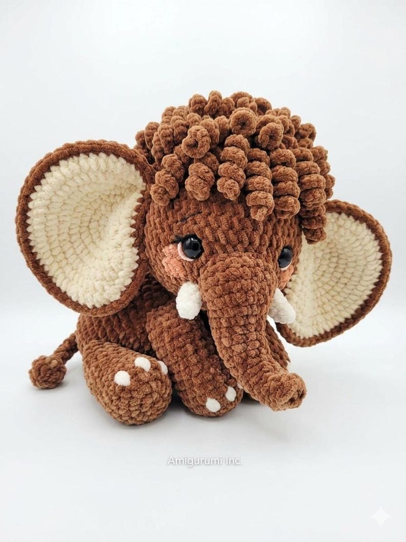 A crochet brown mammoth amigurumi toy sitting on a clean white surface with a soft, curly head mane.