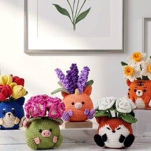 Puede incluir: Una colección de macetas de animales hechas a mano con ganchillo, cada una con flores de ganchillo de colores. Las macetas tienen forma de oso azul, cerdo verde, unicornio naranja, tigre naranja y zorro rojo.