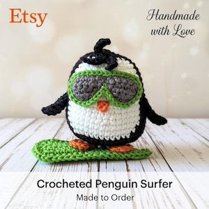 May include: A handmade crocheted penguin surfer with sunglasses. The penguin is black, white, and gray, with a green surfboard. The image includes the text "Etsy" and "Handmade with Love". The caption also includes the text "Crocheted Penguin Surfer Made to Order".