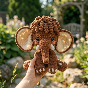 A hand holds a plush brown crochet mammoth toy with large cream-colored ears against a blurred outdoor garden setting.