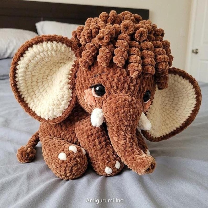 A handmade brown crochet mammoth plush with big floppy ears, white tusks, and a curly mop of hair sits on a gray bed.